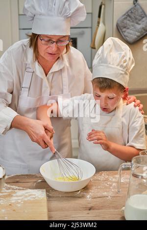 Mutter und Sohn kochen Apfelkuchen in der heimischen Küche. Eine Frau und ein Junge in Küchenhüten und Schürzen kochen mit Gebäck Stockfoto