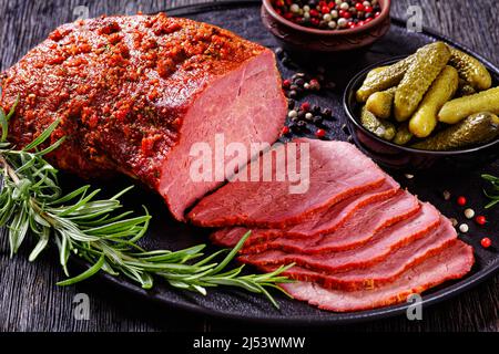 Frisch geschnittenes Rinderpastrami auf schwarzem Teller mit frischem Rosmarin, Thymen und Gurken auf dunklem Tisch, horizontale Ansicht von oben, Nahaufnahme Stockfoto