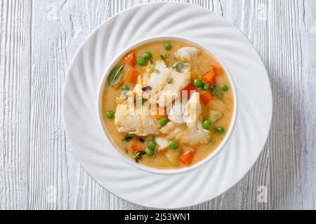 Fischsuppe mit Kabeljau, grünen Erbsen, Karotten, Kartoffeln und Milch auf pflanzlicher Basis, bestreut mit frischem Salbei, Thymian in einer weißen Schüssel auf einem weißen Tisch mit Zutaten Stockfoto