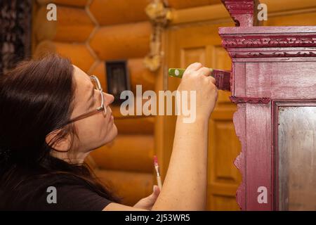 Portrait der alten Frau in Gläsern auf die Aufgabe der Malerei antiken Schrank in roter Farbe mit wenig Pinsel konzentriert. Home Workshop für die Renovierung von Stockfoto