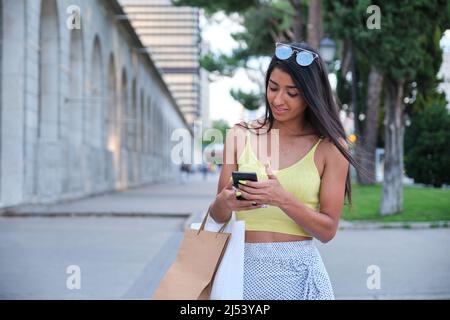 Junge lateinische Frau, die das Telefon benutzt und Einkaufstaschen hält. Stockfoto