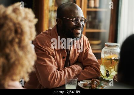 Afrikanischer glücklicher Mann in einer Brille sitzt am Esstisch und lacht mit seiner Familie Stockfoto