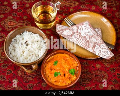 Würziges Huhn Tikka Masala mit geschmortem Reis in 2 Schüssel, Tasse Tee, Teller mit Handtuch und Gabel auf roter Tischdecke. Indische Küche. Stockfoto