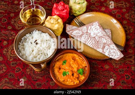 Chicken Tikka Masala mit geschmortem Reis in 2 Schüssel, Tasse Tee, 3 Bambus Gewürzbehälter, Teller mit Handtuch und Gabel auf roter Tischdecke, Nahaufnahme. Indische cui Stockfoto