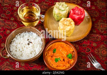 Chicken Tikka Masala, geschmorter Reis, Tasse Tee, 3 Bambus-Gewürzbehälter auf Teller auf rotem orientalischem Tischtuch. Indische Mahlzeit. Stockfoto