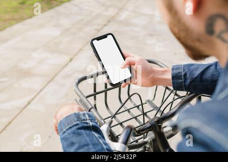 Der junge Mann trägt Denim-Kleidung, lehnt sich an den Lenker des Leihradfahrrads, hält das Smartphone und sucht in den Händen nach einem Telefon, Nahaufnahme. Scrollen Stockfoto