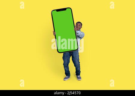 Aufgeregt schwarzer Mann stehen und zeigen großes Smartphone mit leerem grünen Bildschirm über gelben Studio-Hintergrund, Mockup-Bild. Stockfoto