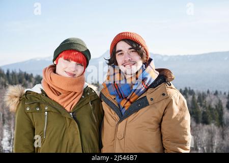 Porträt eines verliebten jungen kaukasischen Mannes und einer verliebten Frau, die am sonnigen Wintertag auf der Bergspitze stehen und die Kamera lächeln Stockfoto