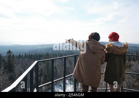 Verliebte junge Männer und Frauen, die irgendwo in den Bergen auf der Aussichtsplattform stehen und die Landschaft am Wintertag betrachten Stockfoto