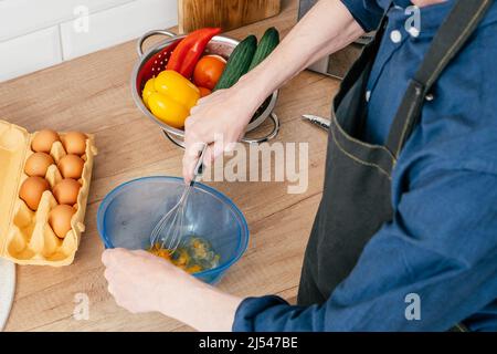 Draufsicht auf einen nicht erkennbaren Mann im blauen Langarm-Hemd, der Eier in einer blauen Kunststoffschüssel mit Drahtbesen auf dem Tisch in der Nähe eines Stahlsieders mit rotem, brüllenden Schrei schlägt Stockfoto