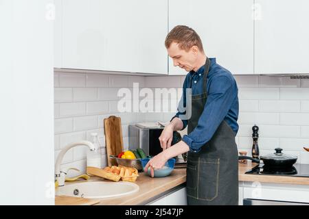 Porträt eines Mannes mittleren Alters mit blauem Langarmhemd und schwarzer Schürze, der in einer blauen Kunststoffschüssel mit Drahtbesen auf dem Tisch in der Küche Eier schlägt. Coo Stockfoto