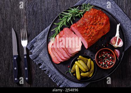 Frisch geschnittenes Rinderpastrami auf schwarzem Teller mit frischem Rosmarin, Thymen und Gurken auf dunklem Tisch, horizontale Ansicht von oben, flach liegend Stockfoto