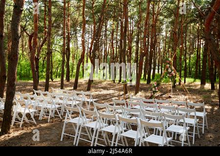 Wunderschöne böhmische Tipi-Bogendekoration auf der Hochzeitsfeier im Freien im Pinienwald mit Kegeln. Stühle, floristische Blumenkompositionen aus Rosen Stockfoto