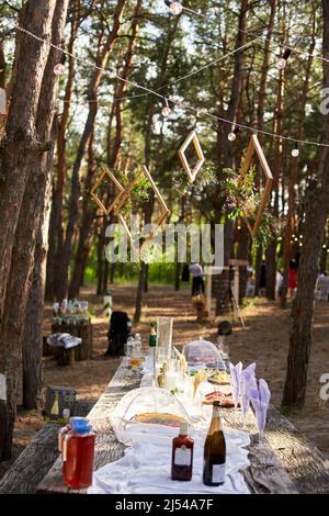 Hochzeitsfeier Bankett im Freien im Wald. Esstisch im Boho-Stil mit Kerzen, weißem Tuch, Blumen, serviert mit Tellern, Geschirr dekoriert Stockfoto