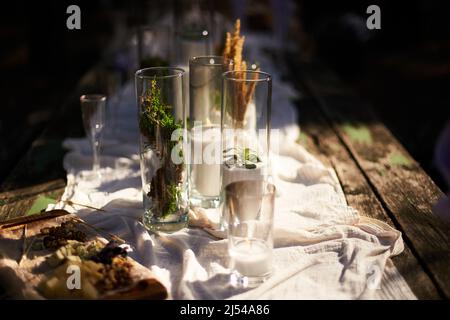 Hochzeitsfeier Bankett im Freien im Wald. Esstisch im Boho-Stil mit Kerzen, weißem Tuch, Blumen, serviert mit Tellern, Geschirr dekoriert Stockfoto