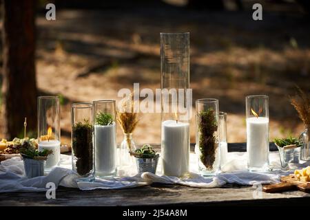 Hochzeitsfeier Bankett im Freien im Wald. Esstisch im Boho-Stil mit Kerzen, weißem Tuch, Blumen, serviert mit Tellern, Geschirr dekoriert Stockfoto