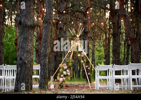 Wunderschöne böhmische Tipi-Bogendekoration auf der Hochzeitsfeier im Freien im Pinienwald mit Kegeln. Stühle, floristische Blumenkompositionen aus Rosen Stockfoto