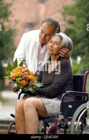 Ältere Paare in der Natur, die Frau sitzt in einem Rollstuhl mit einem Blumenstrauß und der Mann umarmt sie liebevoll Stockfoto