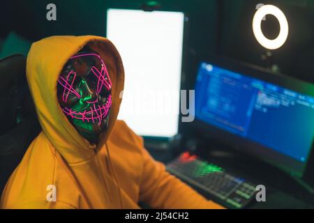 Anonymes Internet-Identitätskonzept. Person in einem gelben Hoodie vor einem Programmiercomputer mit gruselig beleuchteter Neonmaske und Blick auf die Kamera. Hochwertige Fotos Stockfoto