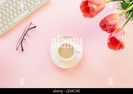 Rosafarbener weiblicher Arbeitsplatz mit Tastatur, Tulpenblüten, Kaffeetasse und Brille. Frühlingsferien, Muttertagskonzept. Draufsicht, flach liegend Stockfoto