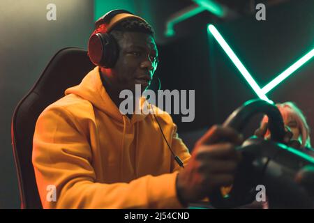 Erwachsener afroamerikanischer Mann mit gelbem Hoodie und großen schwarzen Kopfhörern, der im Spielcafé auf dem Autosimulator rast. Hochwertige Fotos Stockfoto