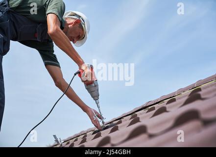 Arbeiter Vorbereitung auf die Installation von Photovoltaik-Solarpanel-System auf dem Dach des Hauses. Mann Installateure arbeiten mit elektrischen Schraubendreher, tragen Uniform und Helm, auf blauem Himmel Hintergrund. Stockfoto