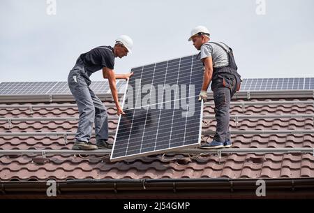 Männer Monteure heben Photovoltaik-Solarmodul auf dem Dach des Hauses. Elektriker in Helmen installieren Solarpanel-System im Freien. Konzept der alternativen und erneuerbaren Energien. Stockfoto