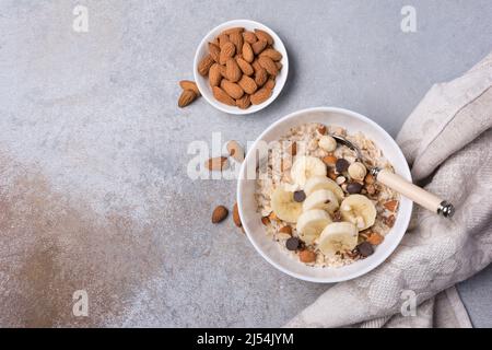 Draufsicht auf Haferflocken mit frischen Bananenscheiben, Mandel-, Haselnuss- und Schokoladentropfen für ein gesundes Frühstück auf grauem Beton-Hintergrund Stockfoto