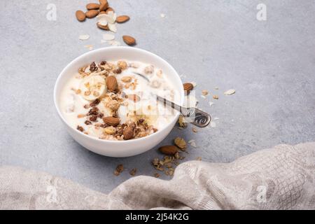 Müsli mit Joghurt, frischen Bananen- und Mandelnüssen für ein gesundes Frühstück auf grauem Betongrund Stockfoto