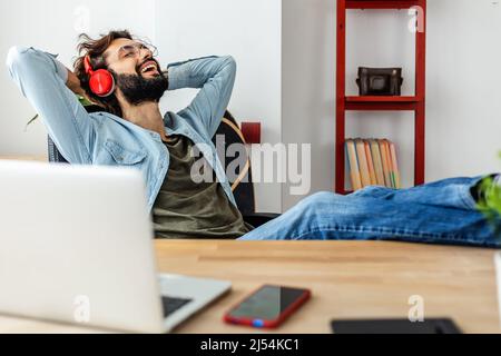 Lächelnder junger, erwachsener Hipster-Mann, der sich am Arbeitsplatz entspannt Stockfoto