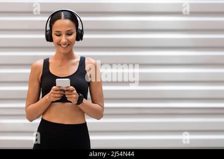 Lächelnde junge arabische Sportlerin in Sportkleidung mit Kopfhörern, die auf grauem Wandhintergrund am Telefon chattet Stockfoto