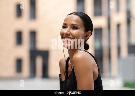 Lächelnde junge arabische Frau Athlet Brünette in Kopfhörern genießt Lieblingsmusik online im Training Stockfoto