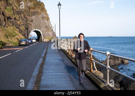 Frau in dunkler Kleidung, die vor den Toren der Drains Bay an der Larne Coast im County Antrim am Meer entlang läuft. Nordirland. Stockfoto