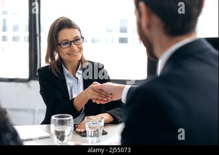 Ein Abkommen zu schließen, die Hände zu schütteln, bestätigt Vereinbarungen. Kaukasische intelligente erfolgreiche Geschäftsfrau, Top-Manager, ceo-Unternehmen, schüttelt die Hände mit männlichen Kollegen in modernen Büro, lächelnd aufeinander Stockfoto
