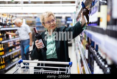 Reife Frau mit Gläsern wählt eine Flasche Wein in der Alkoholabteilung des Supermarkts Stockfoto