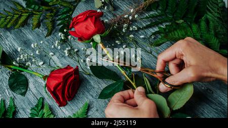 Hochwinkelansicht eines Mannes, der ein Blumenarrangement mit einigen roten Rosen auf einer grauen rustikalen Holzarbeitsfläche macht, in einem Panoramaformat, das als Webbanner verwendet werden kann Stockfoto