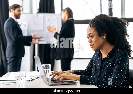 Erfolgreiche positive afroamerikanische Dame bei einem Geschäftstreffen. Ziemlich weibliche Arbeitgeber, Top-Manager, sitzen in einem modernen Büro, arbeiten auf Laptop, entwickeln Start-up, Finanzstrategie, lächelt Stockfoto