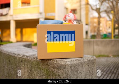 Eine Kiste mit ukrainischer Flagge für die Spende humanitärer Hilfe mit Dingen und Kinderspielzeug für ukrainische Flüchtlinge auf der Straße. Bleiben Sie in der Ukraine. Stockfoto