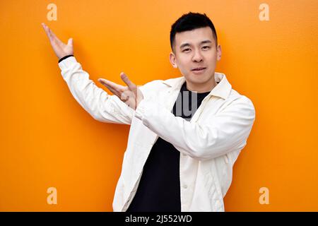Junger Chinese in weißem Hemd auf orangefarbenem Hintergrund, der lächelnd auf die Kamera blickt und mit beiden Händen zur Seite zeigt. Hochwertige Fotos Stockfoto