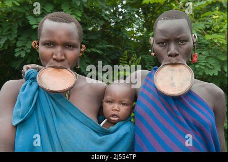 Zwei junge Surma-Frauen mit Lippenbekenntnissen und einem Baby in der Hand, Tulgit, Omo River Valley, Äthiopien Stockfoto