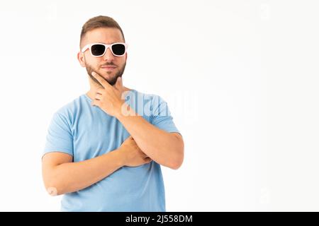 Ein junger Mann, der mit einer Sonnenbrille auf weißem Hintergrund denkt Stockfoto