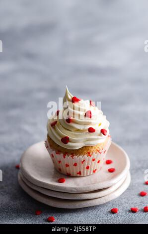 Vanillemuffin mit Buttercreme-Zuckerguss und roten Zuckerherzen auf drei Tellern und grauem Hintergrund mit Kopierraum. Stockfoto