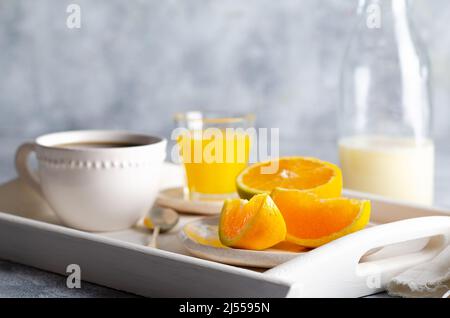 Eine Flasche Milch, eine Tasse Kaffee, Orangensaft und Orangenstücke auf einem weißen Tablett und grauem Hintergrund. Stockfoto