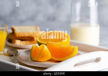 Eine Flasche Milch, ein Glas Orangensaft und Orangenstücke auf einem weißen Tablett und grauem Hintergrund. Stockfoto