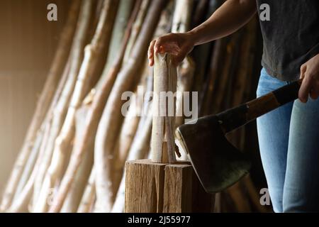 Nahaufnahme der Hand einer Frau mit einem Beil in der einen Hand und einem Stück Holz in der anderen zum Zerkleinern. Baumstämme im Hintergrund Stockfoto