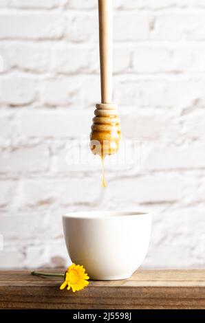 Ein Tropfen Honig, der aus einem Honiglöffel in eine weiße Schüssel tropft, mit einer weißen Ziegelwand im Hintergrund. Stockfoto