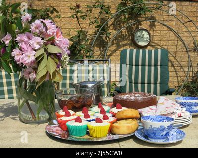 Nachmittagstee in einem englischen Garten: Neben der Teekane, den Tellern und einem Blumenkrug mit Frühlingsblumen wird eine Reihe von Kuchen auf den Tisch gelegt Stockfoto