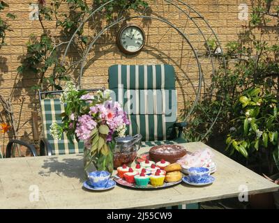 Nachmittagstee in einem englischen Garten: Neben der Teekane, den Tellern und einem Blumenkrug mit Frühlingsblumen wird eine Reihe von Kuchen auf den Tisch gelegt Stockfoto