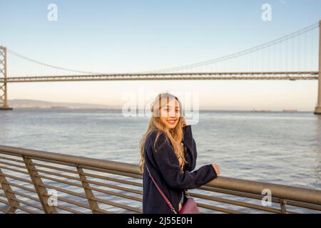 Am späten Nachmittag wurden Halbkörperportraits einer jungen Frau mit der Oakland Bay Bridge und der San Francisco Bay im Hintergrund | Lifestyle Travel in the City Stockfoto