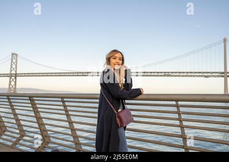 Am späten Nachmittag wurden Halbkörperportraits einer jungen Frau mit der Oakland Bay Bridge und der San Francisco Bay im Hintergrund | Lifestyle Travel in the City Stockfoto